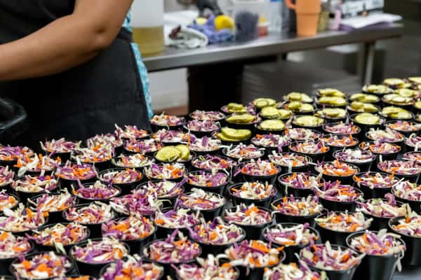 Rows of mini coleslaw cups being prepped in a busy kitchen, each topped with sliced pickles—ideal for Southern BBQ catering and bulk food preparation visuals.