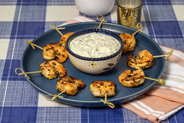 Grilled shrimp skewers arranged around a bowl of creamy herb dip, served on a navy blue platter over a checkered tablecloth—ideal for seafood appetizers or party platters.
