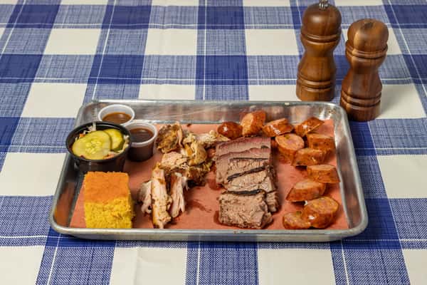 Hearty barbecue tray with sliced brisket, smoked sausage, pulled chicken, cornbread, and house-made pickles with dipping sauces—Southern comfort food served on a metal tray with checkered tablecloth.