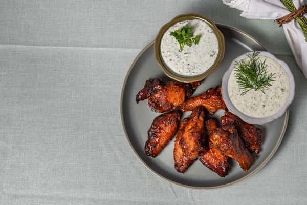 Overhead shot of barbecue chicken wings paired with two cups of creamy ranch dip, garnished with dill and cilantro—perfect for sports bar menus or smoky Southern appetizers.