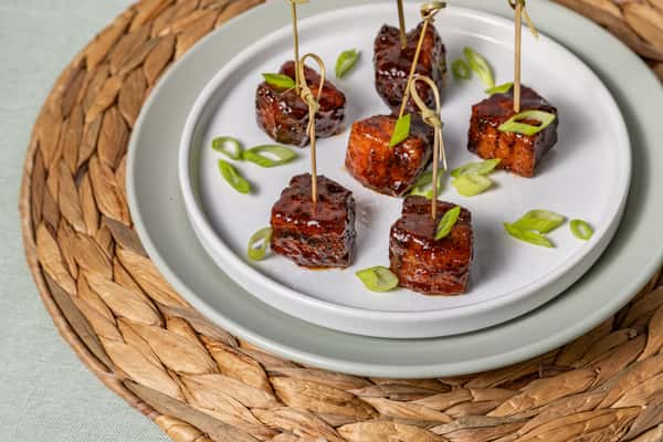 Glazed pork belly burnt ends skewered with cocktail picks and garnished with sliced green onions, elegantly plated on a white dish—perfect for barbecue appetizers or upscale small bites.