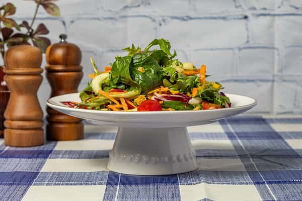Fresh garden salad on a white pedestal plate, featuring cherry tomatoes, shredded carrots, cucumber, and mixed greens—ideal for health-conscious dining or farm-to-table restaurant menus.