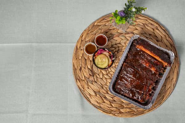BBQ pork ribs served in a foil tray, accompanied by house-made pickles and two dipping sauces, all placed on a woven charger—perfect for showcasing Southern barbecue takeout.