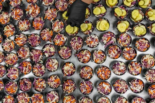 Overhead view of mini slaw cups filled with shredded red and green cabbage, carrots, and topped with pickle chips—perfect for catering or barbecue sides in a commercial kitchen setting.