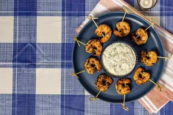 Grilled shrimp skewers arranged in a circular pattern around a bowl of creamy herb dip on a navy plate, set over a blue gingham tablecloth—ideal for Southern seafood appetizers or party platters.