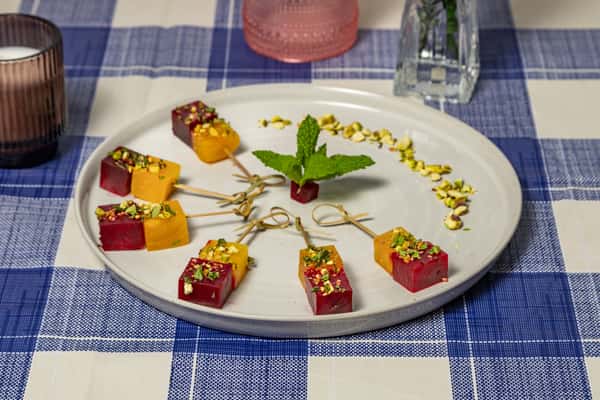 Colorful skewers of beet and golden beet cubes garnished with crushed pistachios and fresh mint, arranged in a circular pattern on a modern white plate—perfect for vegan appetizers or upscale seasonal menus.