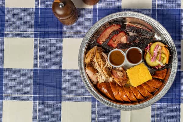 Texas-style barbecue combo platter with sliced brisket, pulled chicken, smoked sausage, cornbread, and pickled cucumber salad, served in a round foil tray—ideal for Southern BBQ catering and family-style meals.