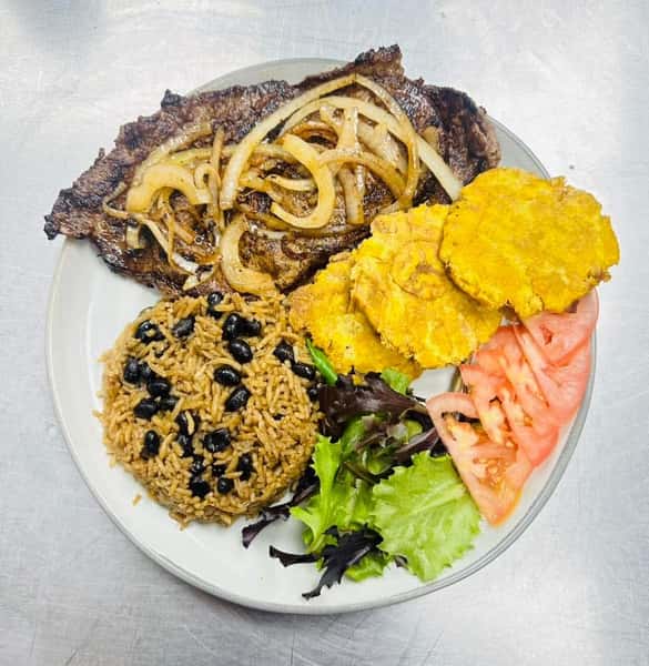 Latin cuisine plate featuring steak with onions, rice with beans, fried plantain and tomato slices.