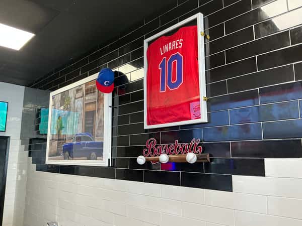 A wall features Cuban baseball memorabilia, including a jersey labeled "LINARES 10," a baseball bat with balls, and a photograph of a blue car. Text: "Baseball." The backdrop is black and white tiled wall.