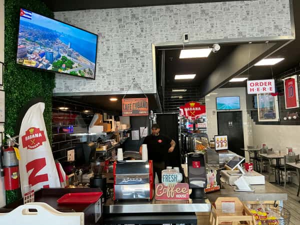 The interior of a Cuban restaurant features the counter, equipment, and staff member, creating an inviting atmosphere. Text includes "CAFE CUBANO," "COFFEE," "FRESH COFFEE HOUSE," and "ORDER HERE."