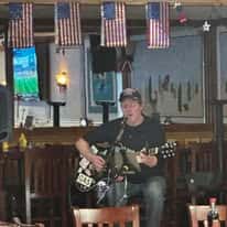 Musician performing live on stage in a casual bar decorated with American flags, playing acoustic guitar and singing into a microphone.