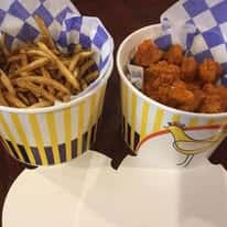 Striped yellow and white takeout cups with checkered paper, one filled with thin fries and the other with spicy popcorn chicken.