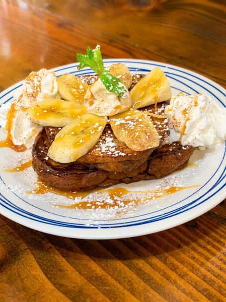 French toast with sliced bananas, whipped cream, powdered sugar, and a mint sprig on a white and blue striped plate.