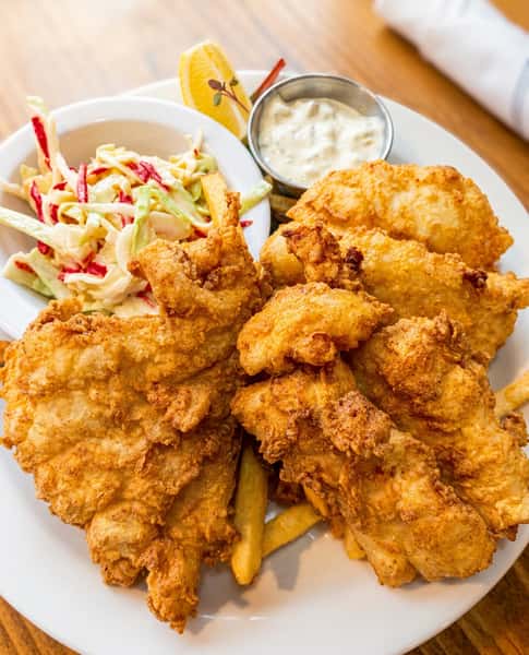 Fried fish and chips with coleslaw, tartar sauce, and a lemon wedge on a white plate.