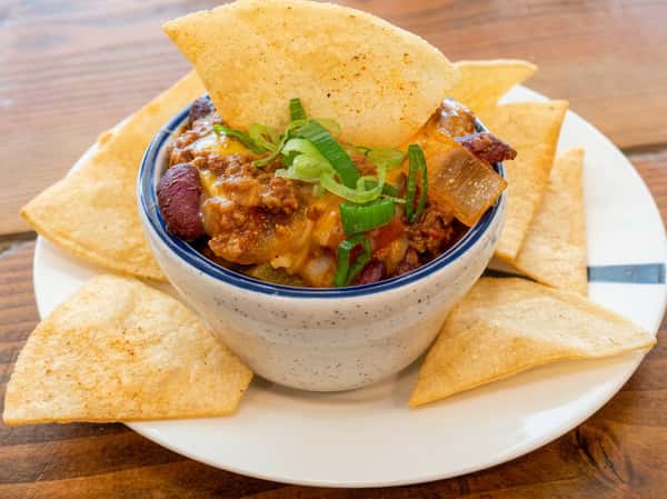 Chili in a speckled white bowl with melted cheese, beans, ground meat, and green onions, surrounded by tortilla chips.