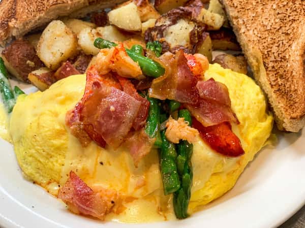 Omelette topped with lobster, bacon, asparagus, and cheese sauce, served with roasted potatoes and toast.
