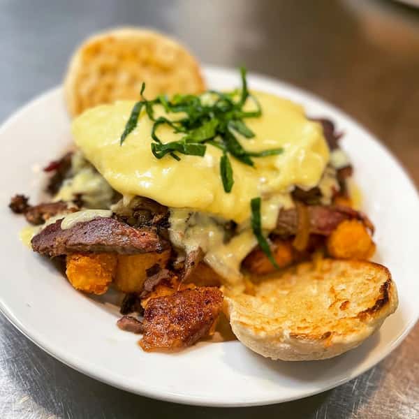 Brisket eggs benedict on a white plate with hollandaise sauce, green garnish, tater tots, and biscuit halves.