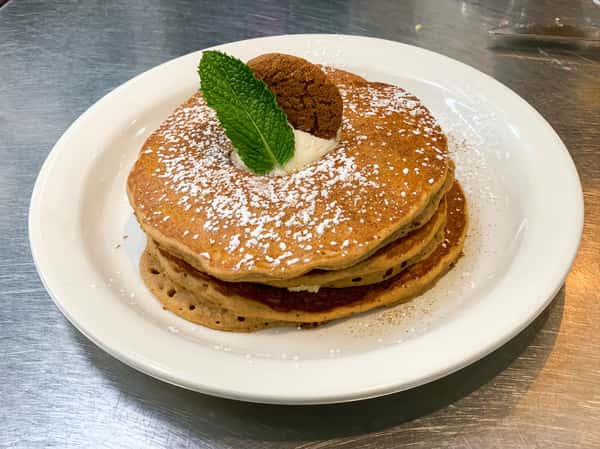 Stack of pancakes with powdered sugar, a dollop of white cream, a mint leaf, and a cookie on a white plate.