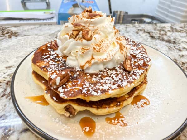 Pancakes topped with whipped cream, pecans, powdered sugar, and syrup on a white plate.