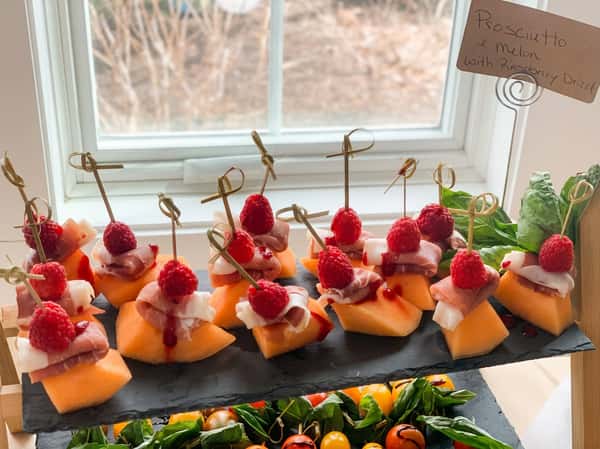 Prosciutto e melon bites with raspberries and a raspberry drizzle on a slate tray.