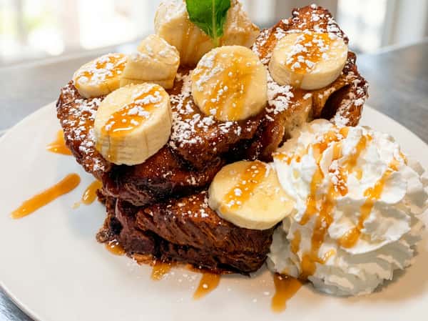 French toast stack with sliced bananas, whipped cream, powdered sugar, and caramel drizzle.
