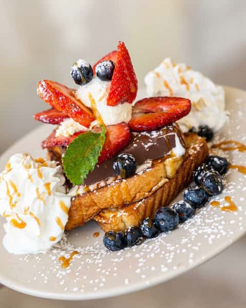 French toast topped with strawberries, blueberries, whipped cream, a chocolate bar, mint, and powdered sugar.