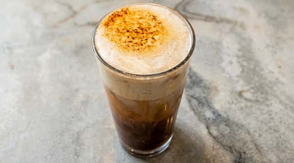Cafe Remedy | Glass of latte with foamy milk and a sprinkling of spices on top, sitting on a marbled surface.