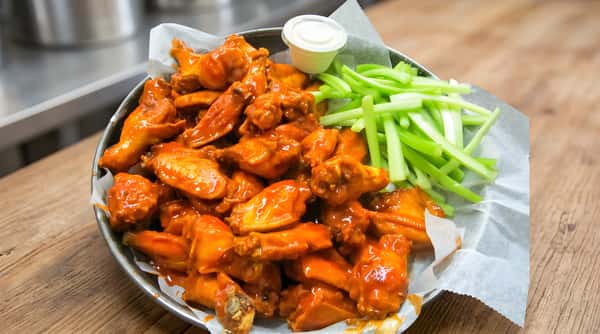 Basket of buffalo chicken wings with celery sticks and a cup of dipping sauce, on white parchment paper.