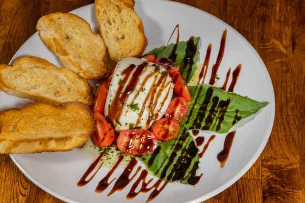 Fresh Caprese salad with mozzarella, cherry tomatoes, and basil leaves drizzled in balsamic glaze, served with toasted crostini on a white plate atop a wooden surface.
