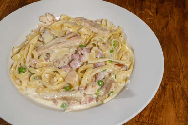 Creamy fettuccine Alfredo tossed with grilled chicken, diced ham, green peas, and a rich garlic Parmesan sauce, served on a white plate over a wooden table.