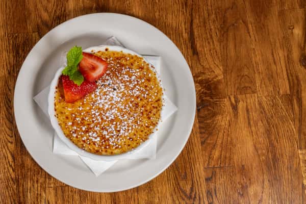 Top‑view of crème brûlée with a golden torch‑caramelized top, garnished with fresh strawberries and mint on a white plate.