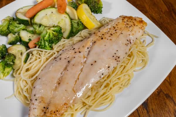 Grilled fillet of fish placed over angel hair pasta, served with steamed mixed vegetables on a white plate.