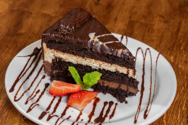 Slice of rich chocolate layer cake with chocolate frosting and creamy filling, drizzled with chocolate sauce, and topped with strawberries and mint on a white plate.