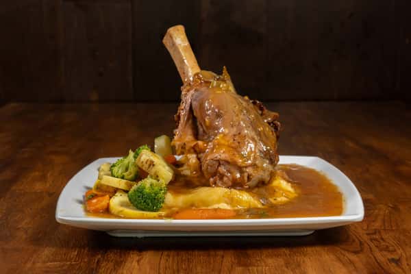 Slow-braised lamb shank in savory brown gravy, served over mashed potatoes with a medley of steamed vegetables on a square white plate.
