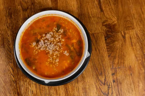 Hearty Italian sausage soup with ground meat, spinach, and pasta in a tomato-based broth, served in a white bowl over a black plate.