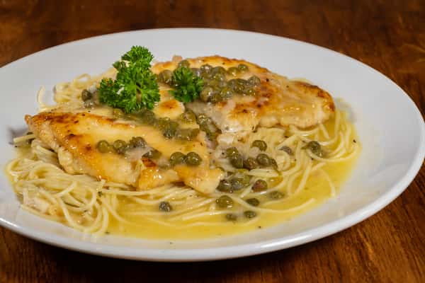 Tender chicken piccata topped with capers and lemon butter sauce, served over angel hair pasta and garnished with parsley on a white plate over a wooden table.