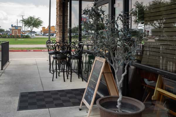 Restaurant entrance featuring a chalkboard sign, potted plant, high top tables, and glass window walls.
