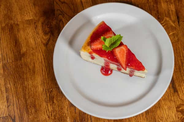 Slice of strawberry cheesecake topped with fresh strawberry slices and a mint leaf, served on a white plate.