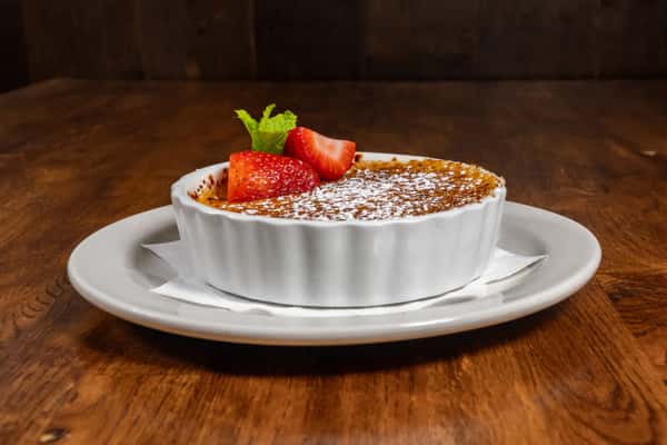 Silky crème brûlée with a caramelized sugar crust, garnished with sliced strawberries and fresh mint, served in a white ramekin over a plate.