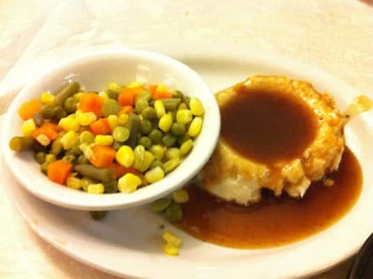 mashed potatoes with gravy on a plate with a small white bowl of corn, peas and carrots.