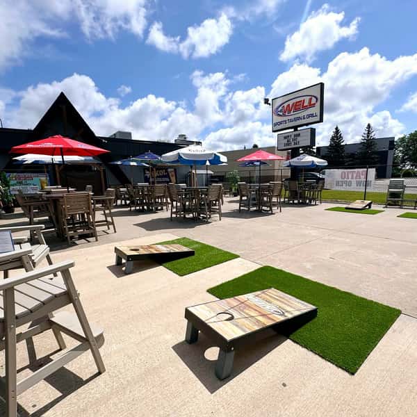 The outdoor patio area with cornhole games, tables, chairs and umbrellas at The Well Sports Tavern and Grill