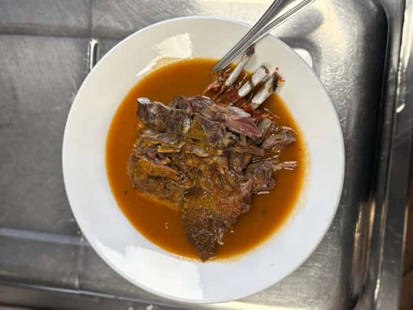Plate of birria featuring tender shredded beef ribs in a rich, red consommé, served in a white bowl with a fork and spoon.