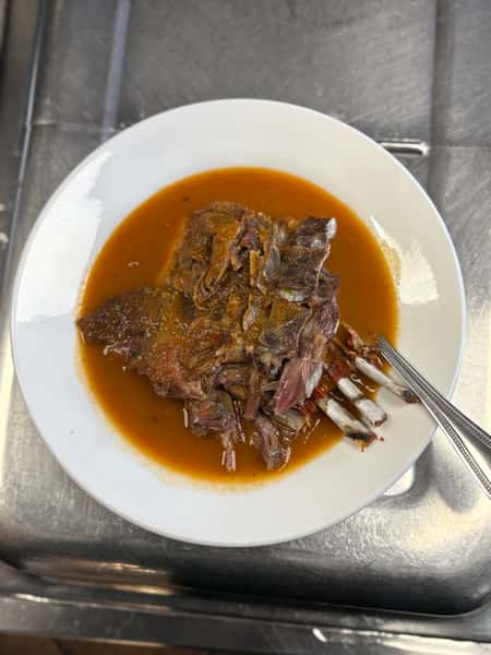 Plate of birria featuring tender shredded beef ribs in a rich, red consommé, served in a white bowl with a fork and spoon.