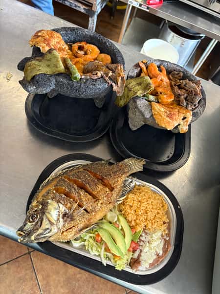Two molcajetes filled with grilled shrimp, meat, cactus, and cheese alongside a plate with fried whole fish, rice, refried beans, and salad.