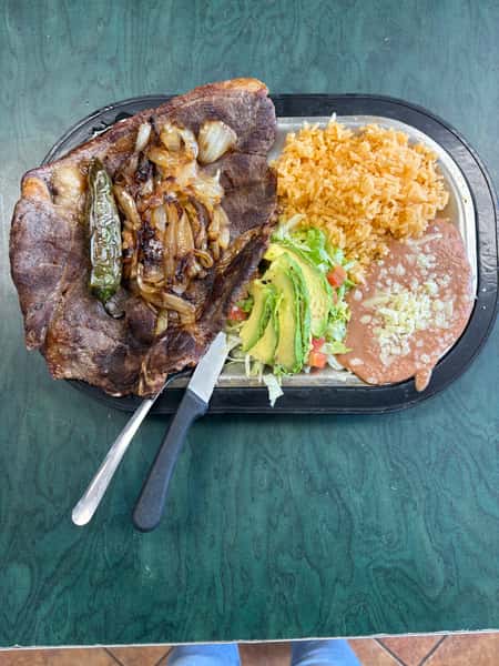 Grilled beef steak with sautéed onions and jalapeño, served with Mexican Grilled beef steak with sautéed onions and jalapeño, served with Mexican rice, refried beans, and avocado salad on a metal plate.rice, refried beans, and avocado salad on a metal plate.