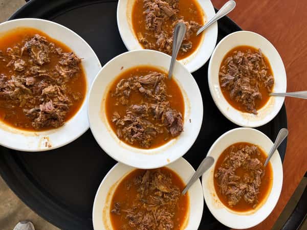 Six white bowls filled with tender birria beef in a rich red broth, each served with a metal spoon on a black tray.