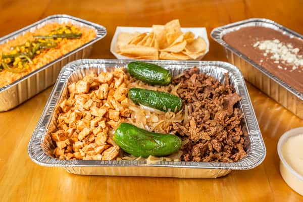 Tray of chicken and beef fajitas with grilled green peppers, with rice, beans, and chips behind it.