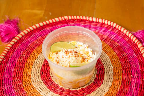 Angled view of a corn cup topped with cheese and seasoning, served with lime on a woven mat.