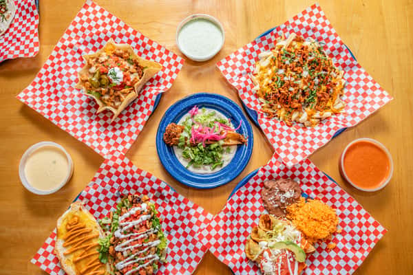 Overhead spread of tacos, loaded dishes, flautas, and dips arranged around a blue plate.
