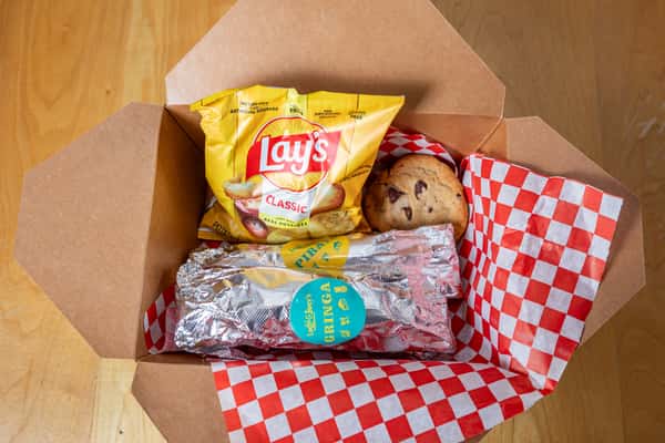 Open cardboard lunch box with a foil-wrapped item, a bag of potato chips, and a cookie.